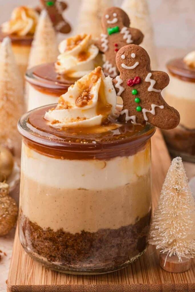 Layered gingerbread trifle in a glass jar with a spiced cake base, creamy filling, whipped topping, and caramel drizzle, garnished with a smiling gingerbread cookie. A festive no-bake Christmas dessert surrounded by mini white bottle brush trees.