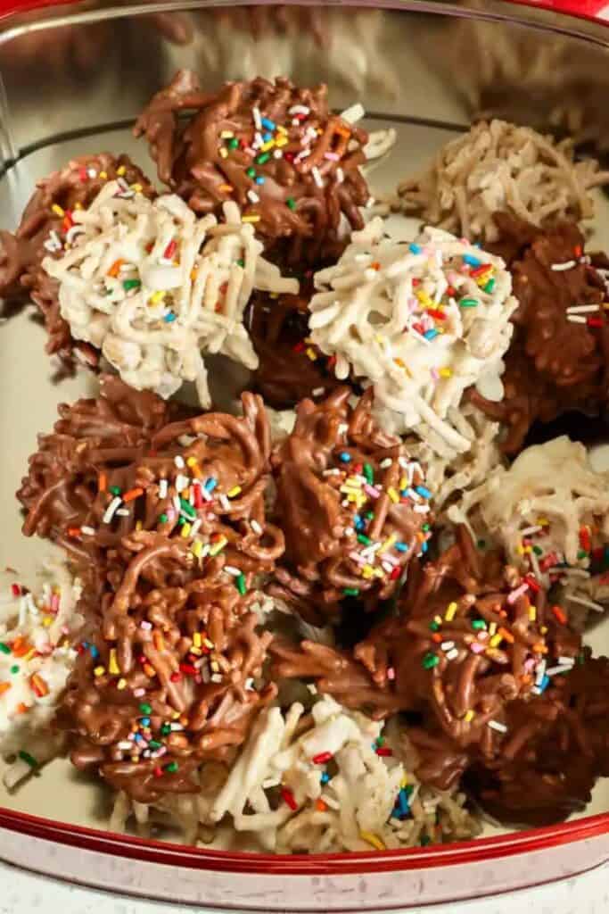 Assorted no-bake chocolate haystack cookies made with chow mein noodles coated in milk or white chocolate, topped with colorful rainbow sprinkles displayed in a festive holiday tin.