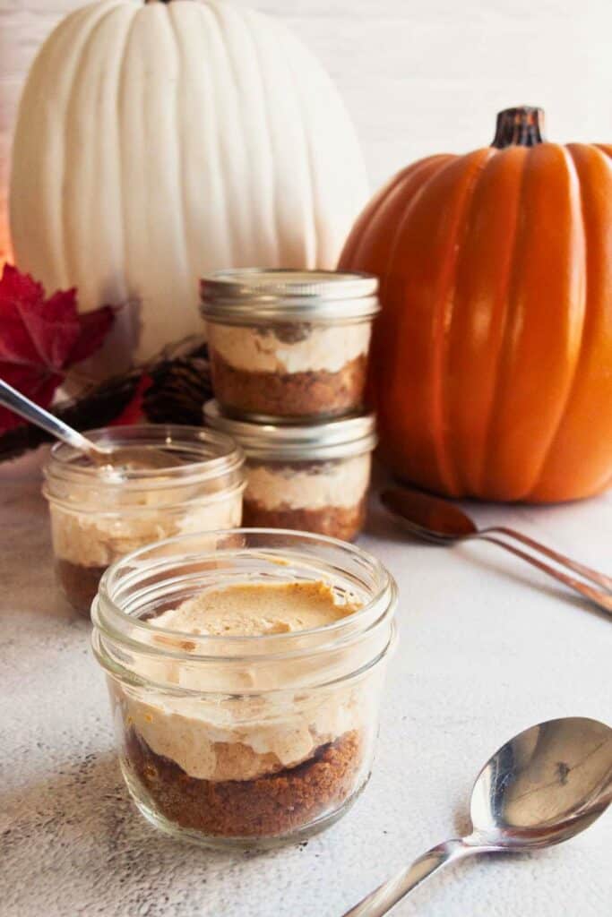 No bake mini pumpkin cheesecake in a glass jar with a crumbly brown crust and creamy spiced pumpkin filling. Several individual cheesecake cups sit beside orange and white pumpkins for a festive dessert display.