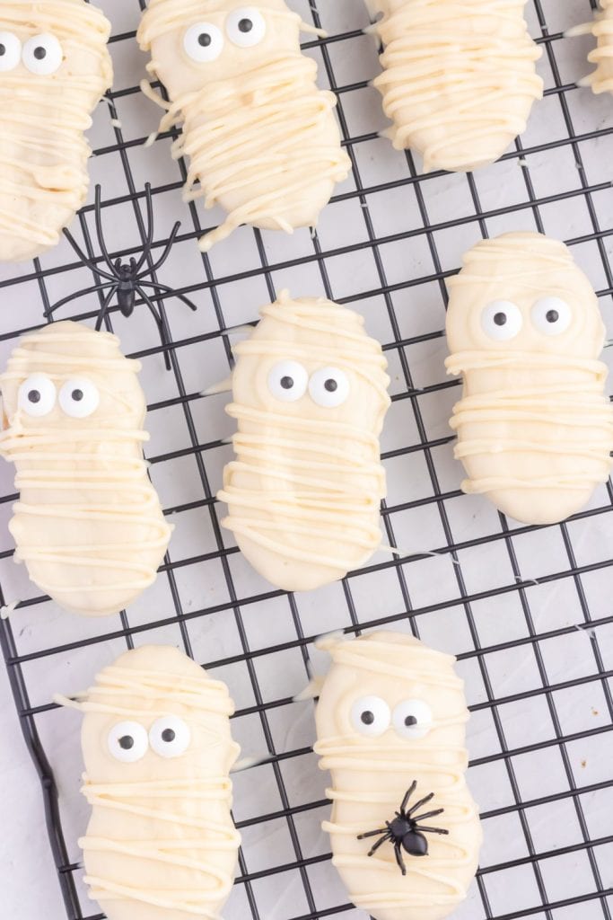Halloween mummy cookies made with oval-shaped Nutter Butter cookies coated in white chocolate and drizzled with more white chocolate to mimic mummy wrappings, topped with candy eyes. Cookies are displayed on a black cooling rack with a few small plastic spiders for decoration.