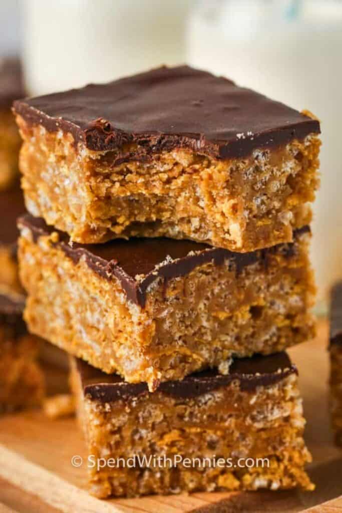 A stack of thick No Bake Peanut Butter Bars with a chewy peanut butter cereal base and a smooth chocolate layer on top. One bar has a bite missing to reveal the dense crunchy texture inside.