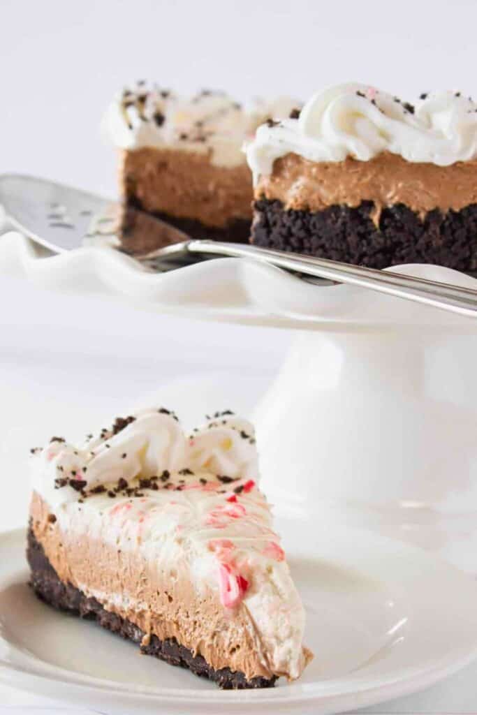 Slice of no bake chocolate peppermint cheesecake with a crushed chocolate cookie crust, creamy chocolate and peppermint layers, and a swirl of whipped topping sprinkled with crushed candy canes. Served on a white plate for a festive holiday dessert.