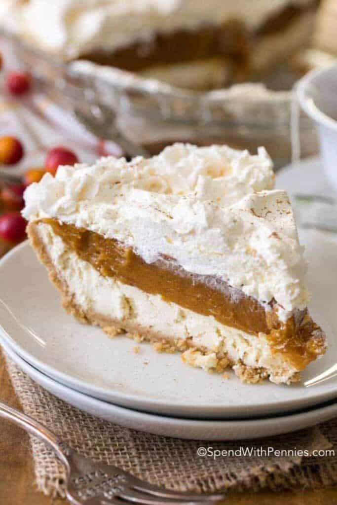 A slice of layered no bake pumpkin dessert sits on a white plate with a crumbly graham cracker crust, creamy cheesecake filling, spiced pumpkin layer, and fluffy whipped topping. The distinct layers create a rich fall dessert with pumpkin and cream in every slice.