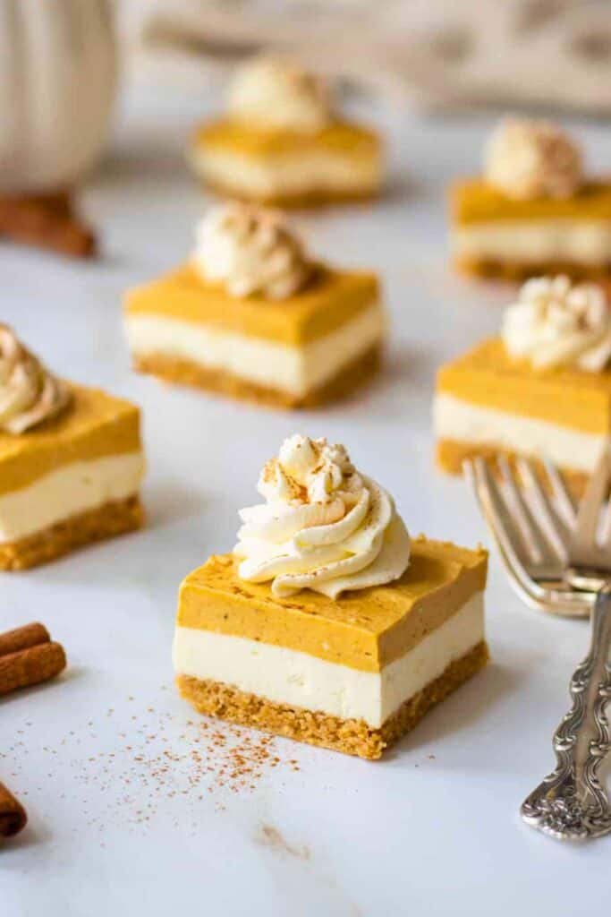No bake pumpkin cheesecake bars with a crumbly golden crust and distinct layers of creamy cheesecake and spiced pumpkin filling. Each square is topped with a swirl of whipped cream and a sprinkle of cinnamon.