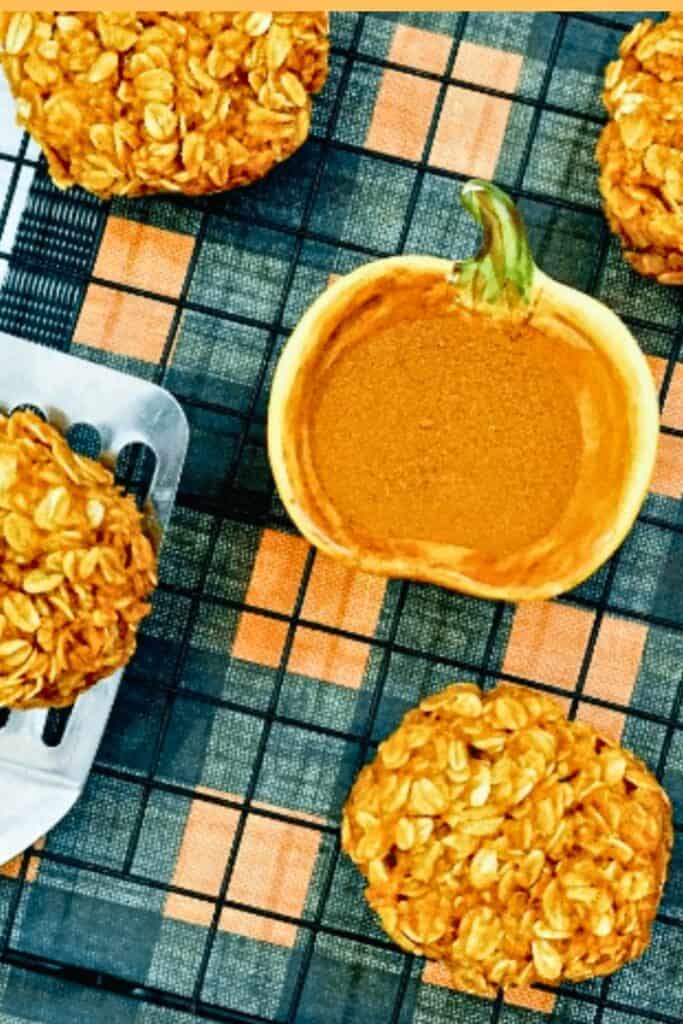 No bake pumpkin cookies made with rolled oats arranged around a small pumpkin shaped bowl filled with creamy pumpkin dip. The golden orange cookies are displayed on a black and orange plaid cloth for a festive fall treat.