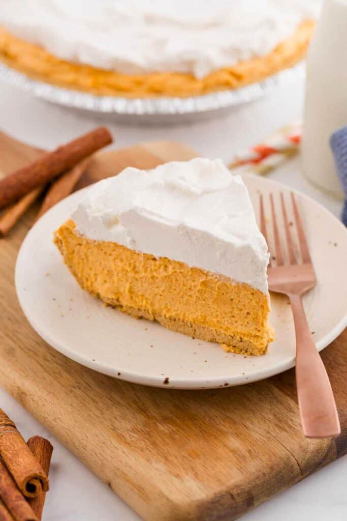 A slice of no bake pumpkin pie with a creamy orange pumpkin filling, crumbly graham cracker crust, and thick whipped topping sits on a white plate. Cinnamon sticks and the remaining pie create a cozy fall setting in the background.