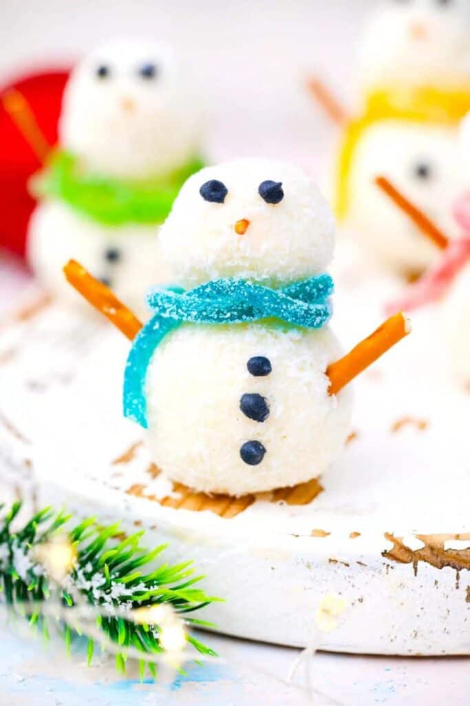 Adorable no bake Snowman truffles made from coconut-covered balls stacked to resemble snowmen, with candy eyes, blue gummy scarves, pretzel stick arms, and chocolate button details.