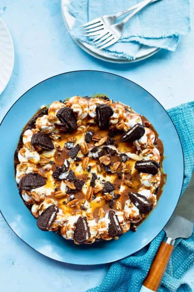 No Bake Turtle Pie topped with swirls of whipped cream, caramel sauce, pecans, and broken chocolate sandwich cookies. The rich chocolate pie is served on a blue platter with extra cookie crumbs and pecans scattered across the top.