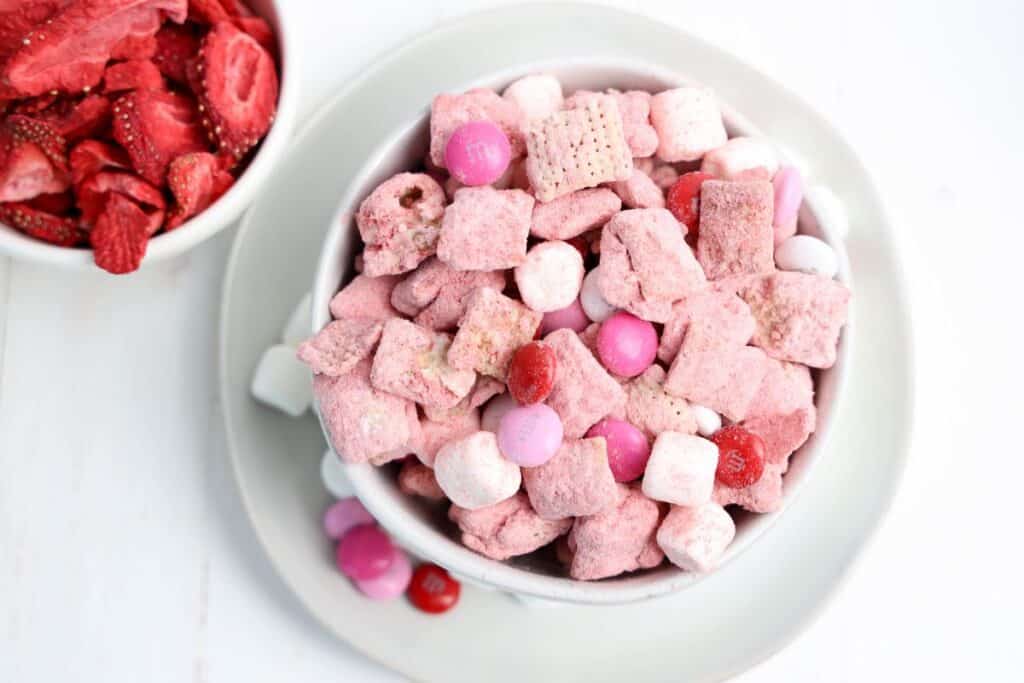 An overhead view of a medium sized bowl of Strawberry Puppy Chow snack mix with red, white and pink M&Ms and mini marshmallows.