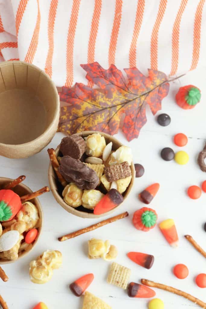 Overhead view of no bake fall snack mixes served in small brown cups, filled with caramel popcorn, Chex cereal, candy corn, candy pumpkins, chocolate-covered pretzels, mini peanut butter cups, and pretzel sticks, with scattered mix ingredients and fall-themed decor on a white surface.