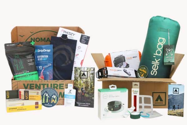 Two open Nomadik subscription boxes filled with outdoor adventure gear, including an emergency poncho, hydration packets, fire starter, compact cook set, camping utensils, a green sleeping bag labeled “sek•bag,” a headlamp, and weather-resistant phone pouch. Items are arranged to highlight their utility for hiking, camping, and survival preparedness.