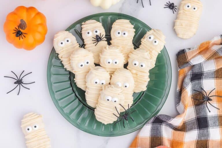 A green plate filled with Halloween-themed Nutter Butter mummy cookies, each coated in white chocolate and decorated with candy eyes and piped white chocolate "bandages." The cookies are surrounded by fake spiders, mini pumpkins, and a plaid orange-and-black napkin for a festive spooky Halloween vibe.