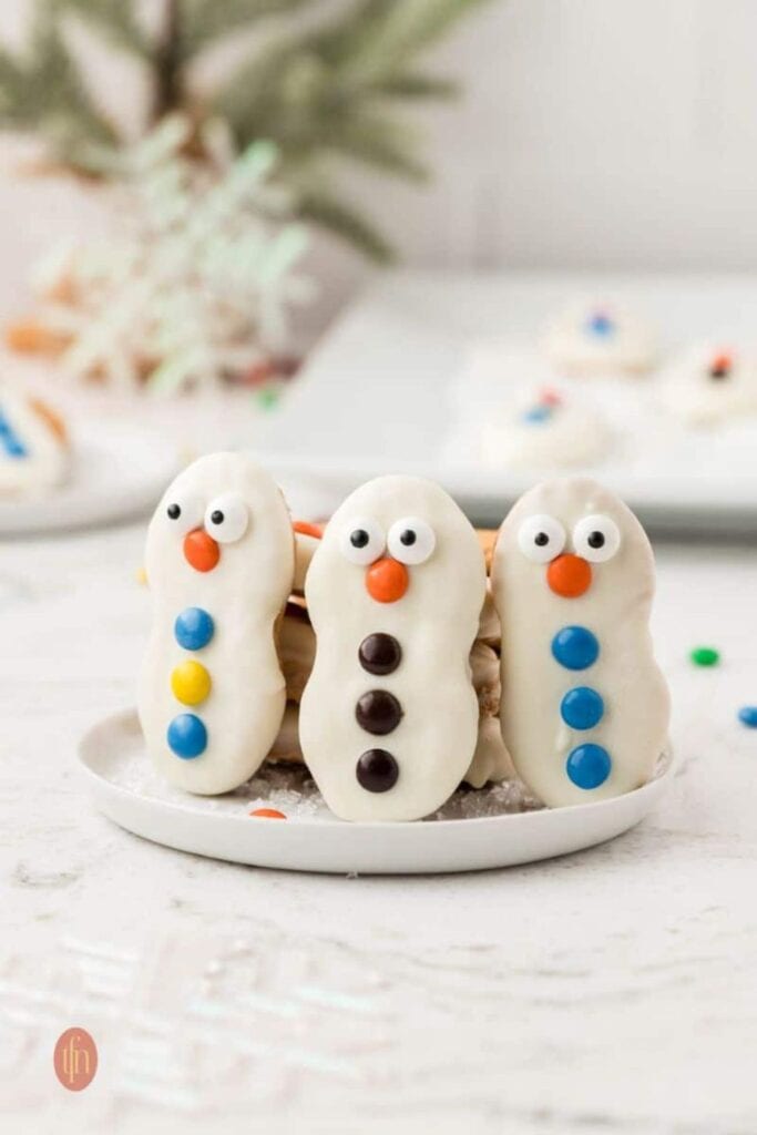 Nutter Butter Snowman cookies dipped in white chocolate and decorated with candy eyes, orange noses, and colorful candy buttons, arranged upright on a plate.