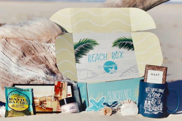 Oceanista Beach Box displayed on sandy beach with driftwood, featuring a tropical-themed open box labeled “Beach Box by Oceanista,” bath salts, body butter, a starfish-shaped soap, a seashell candle, a chocolate bar, and a blue mug reading “All you need is love & the beach.” The setup evokes a relaxing coastal getaway.