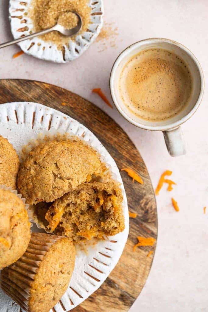 Top-down view of moist, golden-brown carrot cake muffins on a rustic white and wooden plate, with one muffin split open to reveal shreds of carrot inside. A cup of espresso and a dish of raw sugar sit nearby, highlighting this easy, one-bowl recipe as a wholesome snack or breakfast treat.