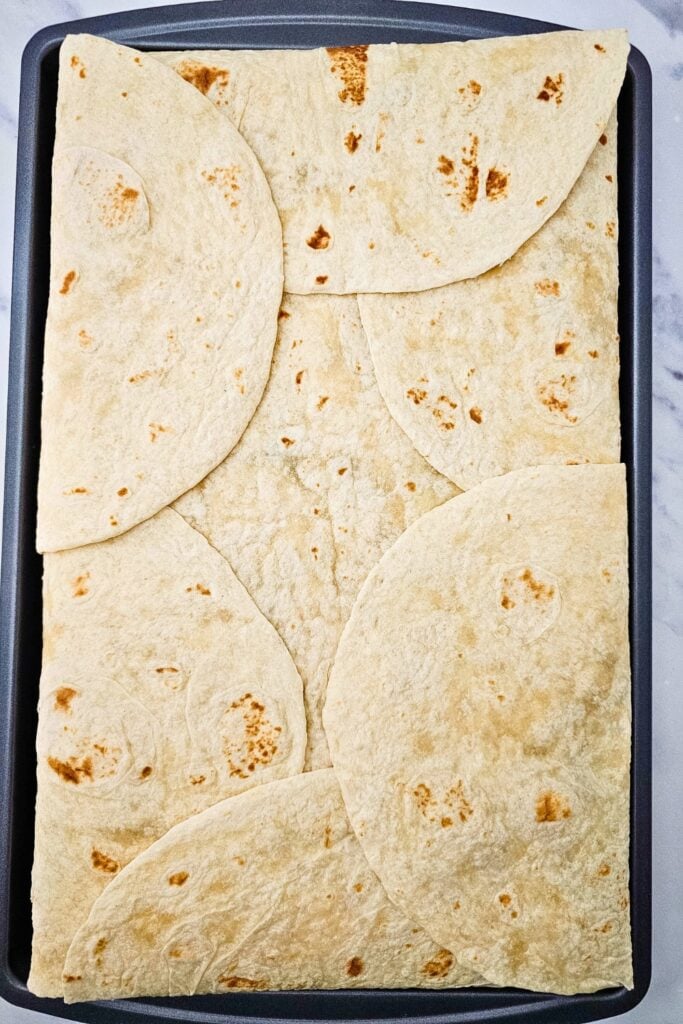 Top-down view of a full sheet pan covered with overlapping flour tortillas, sealing in the cheesy, beef and veggie quesadilla filling underneath. This is the final step before baking, ensuring a crispy, golden top for this easy oven quesadilla recipe.