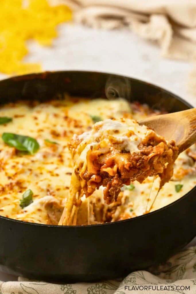One pan lasagna skillet with layers of pasta, seasoned ground beef, tomato sauce, and melted mozzarella bubbling in a cast iron pan. A wooden spoon lifts a cheesy portion from the skillet with melted cheese stretching from the pan.