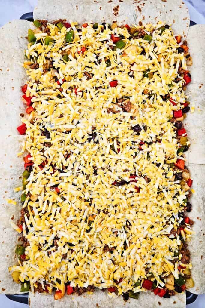 One sheet pan loaded with flour tortillas, filled with a colorful mix of seasoned ground beef, black beans, red and green peppers, onions, and green chilies, all topped with shredded Colby Jack cheese. The filling is evenly spread and ready to be baked into easy, cheesy one pan quesadillas.