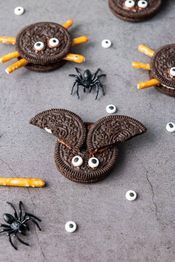 Halloween-themed Oreo cookie creatures on a gray surface, including spider cookies made with pretzel stick legs and candy eyes, and a bat cookie with split Oreo wings. Scattered candy eyes and small plastic spiders add a playful, spooky touch.