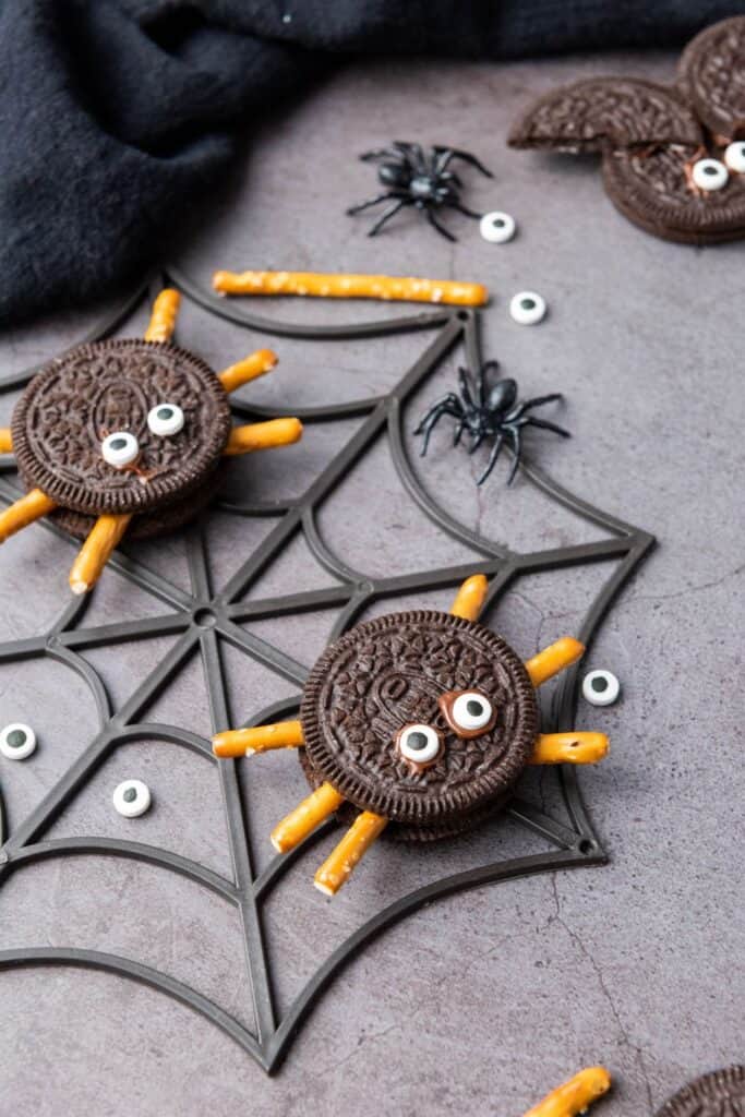 Two oreo cookie spiders with candy eyes and pretzel stick legs on a plastic spider web.