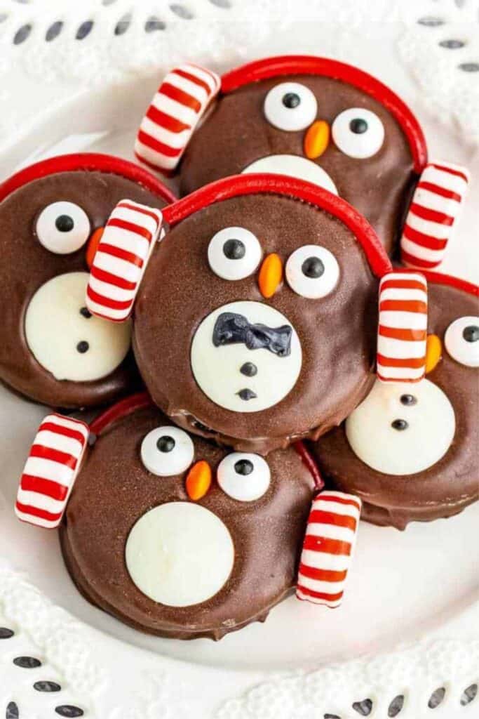 Adorably decorated no-bake Christmas cookies shaped like penguins feature chocolate-coated sandwich cookies with candy eyes, orange sprinkle beaks, white candy bellies, and red-striped candy earmuffs.