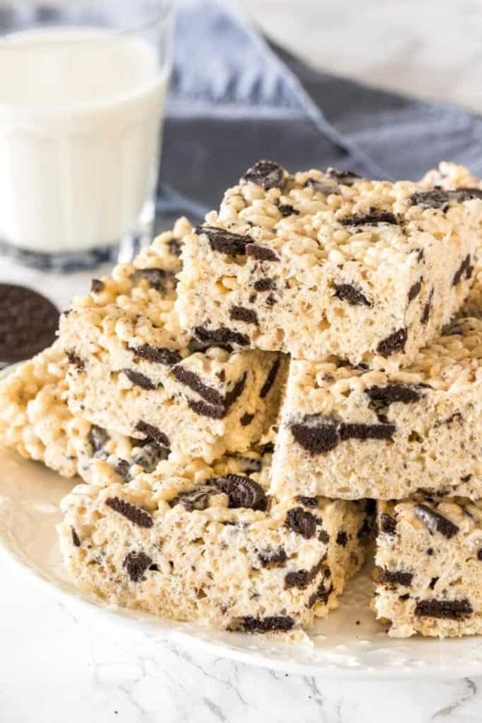 Oreo Rice Krispie squares made with chunks of chocolate sandwich cookies mixed into marshmallow-coated cereal, stacked on a plate with a glass of milk in the background.