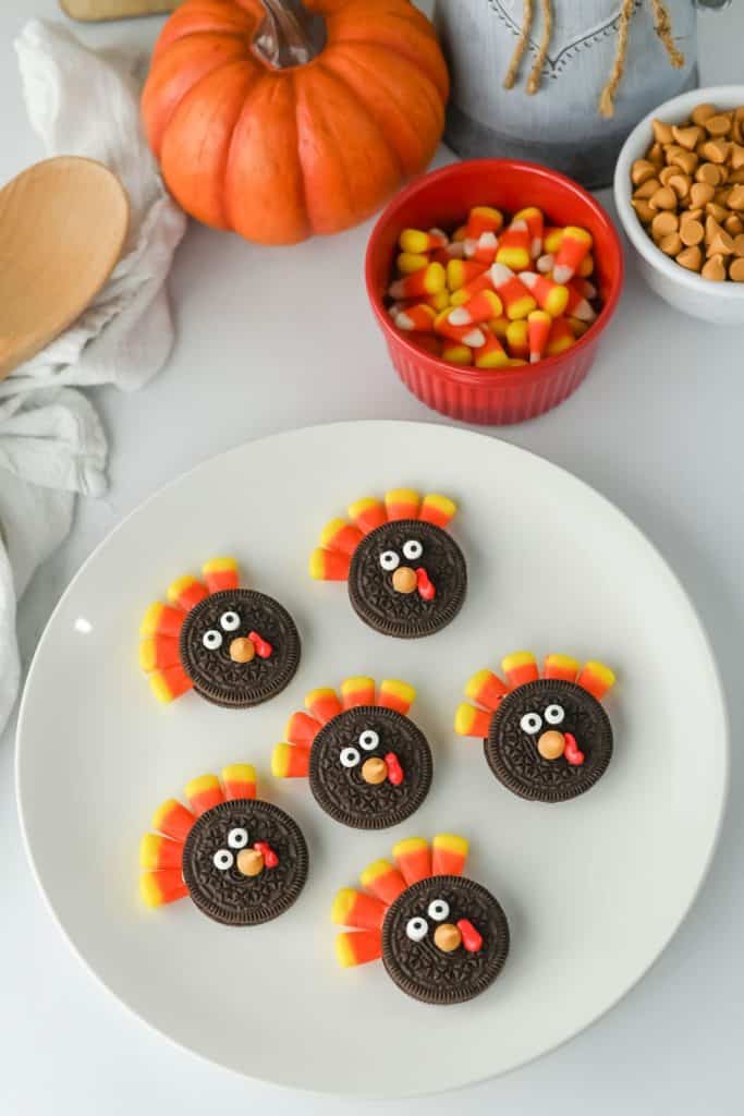 Plate of Oreo Turkey Cookies made with sandwich cookies decorated to look like turkeys, using candy eyes, red icing for the wattle, and candy corn feathers. Surrounding the plate are fall-themed elements like a small pumpkin, a red bowl of candy corn, and a bowl of butterscotch chips.
