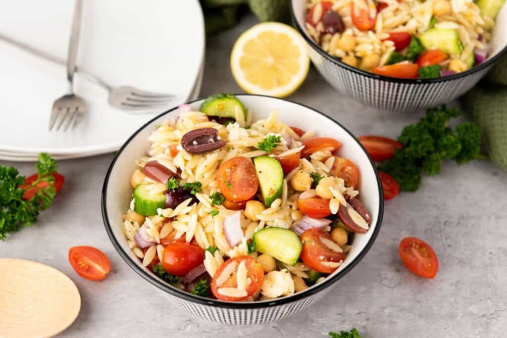 Two bowls of Orzo Mediterranean pasta salad with cherry tomatoes, kalamata olives, cucumbers, and chickpeas.