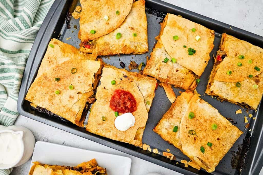 Overhead shot of a sheet pan filled with baked quesadillas fresh from the oven, each piece golden brown and topped with sliced green onions, with one piece garnished with salsa and sour cream. The crispy tortillas reveal a cheesy, savory filling ideal for a quick oven-baked meal.