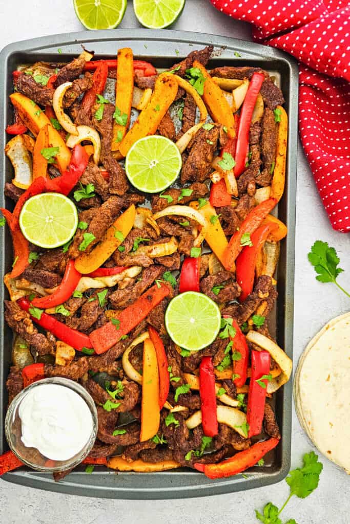 Overhead view of oven baked fajitas with seasoned steak strips. Red and orange bell peppers and onions are arranged on a sheet pan with fresh cilantro and lime halves. A bowl of sour cream and a stack of flour tortillas sit alongside the pan.