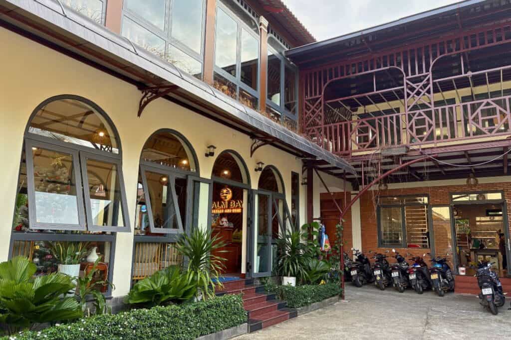 Exterior of the Palm Bay Resort office and lobby area with arched windows, tropical plants, and a row of parked motorbikes along the entrance. A red wooden balcony structure spans the upper level, and festive decor can be seen through the windows.