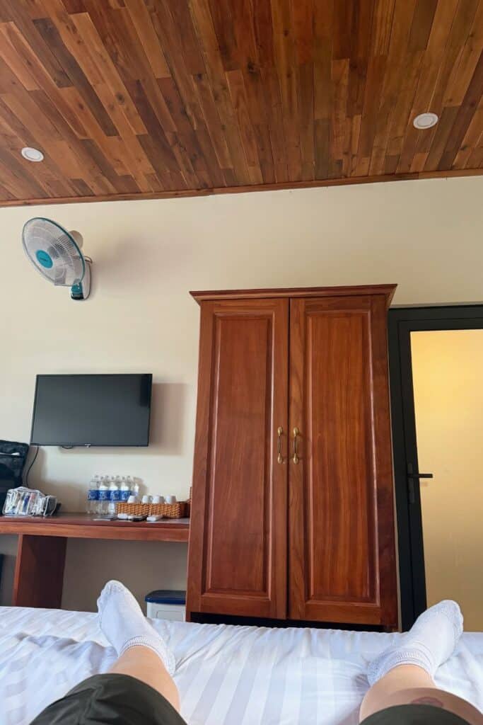 Relaxing inside a family friendly resort room at Palm Bay Resort, Phu Quoc, with a view of a wooden wardrobe, flat-screen TV, wall fan, and complimentary bottled water on a desk. The photo is taken from the perspective of someone lying on a bed with white-striped sheets and wearing white socks.