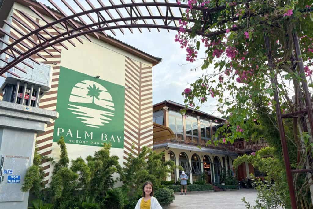 Entrance to Palm Bay Resort in Phu Quoc, Vietnam, featuring a large green logo with a palm tree and ocean graphic on the wall. A smiling young girl stands in front of tropical greenery, while a man checks his phone near the arched entrance framed by blooming pink bougainvillea.