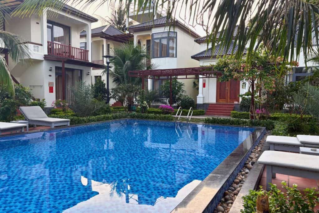 Quiet courtyard pool area at Palm Bay Resort in Phu Quoc, surrounded by white villa-style rooms, lush gardens, and cushioned lounge chairs. The blue-tiled swimming pool reflects the palm trees and sky, offering a serene setting for a relaxing stay.