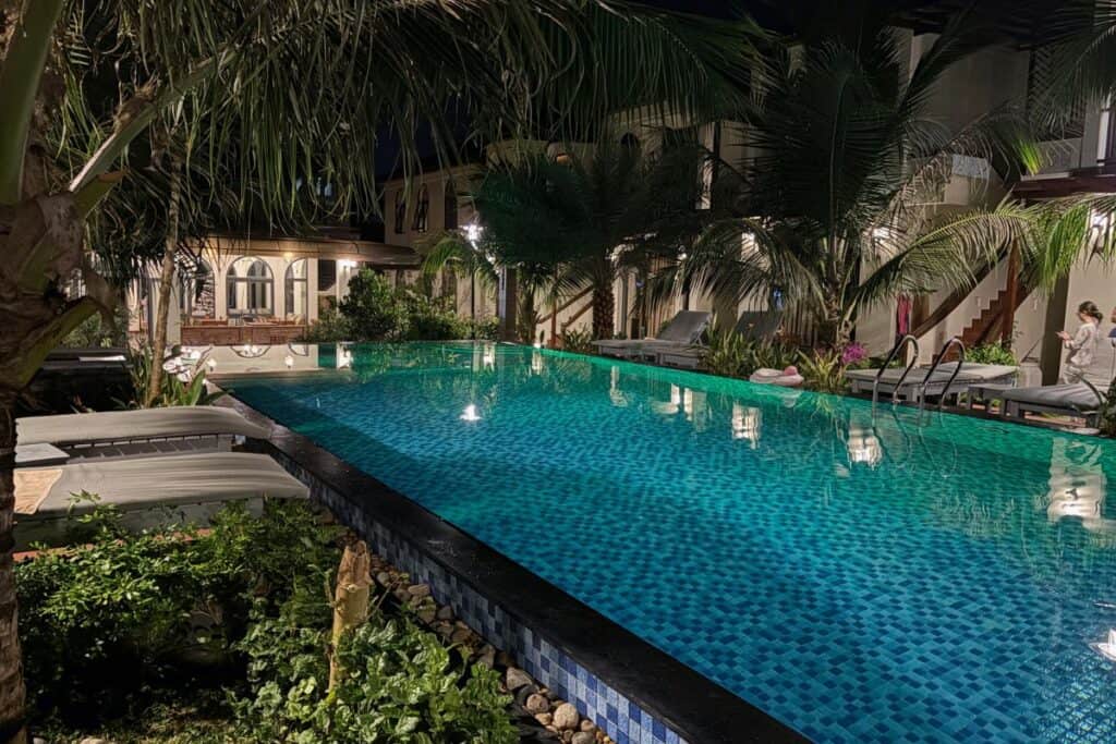 Peaceful night view of the swimming pool at Palm Bay Resort, a family friendly resort in Vietnam, surrounded by palm trees, cushioned lounge chairs, and warmly lit villa-style rooms, creating a relaxing tropical atmosphere after dark.