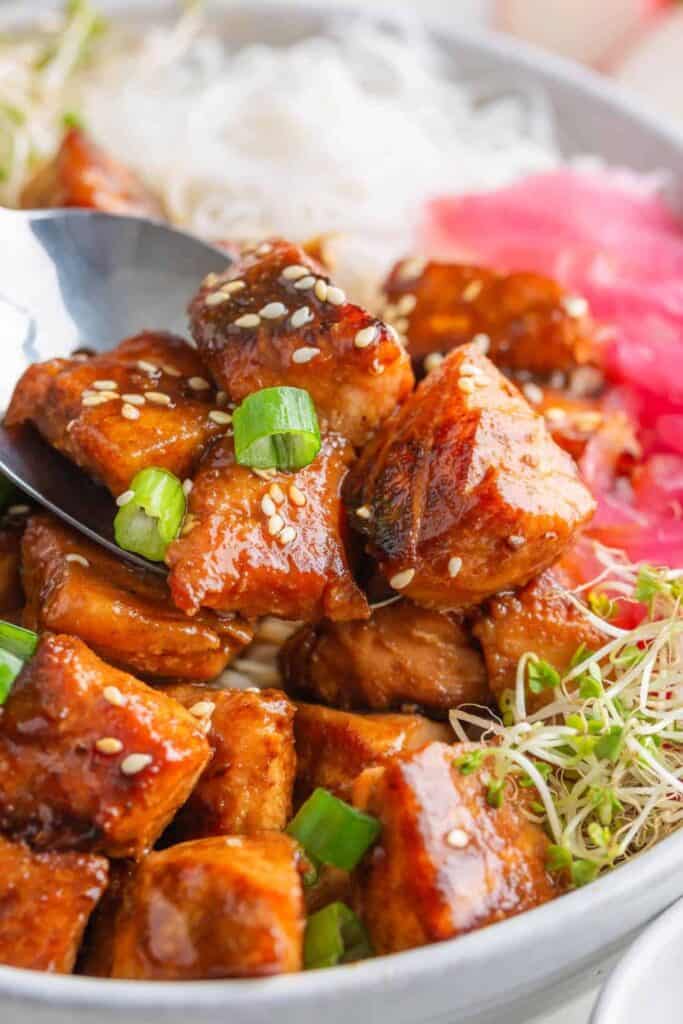 Salmon bites glazed in a sticky sauce and topped with sesame seeds and sliced green onions served over rice. A spoon lifts several salmon pieces from the bowl with pickled ginger and fresh sprouts on the side highlighting this quick dinner recipe.