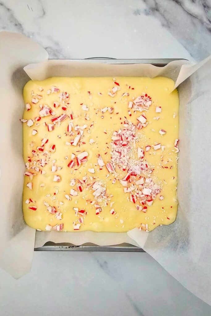 White chocolate fudge mixture in a parchment-lined pan topped with a generous sprinkle of crushed candy canes. This festive layer is ready to chill and set into peppermint holiday fudge.