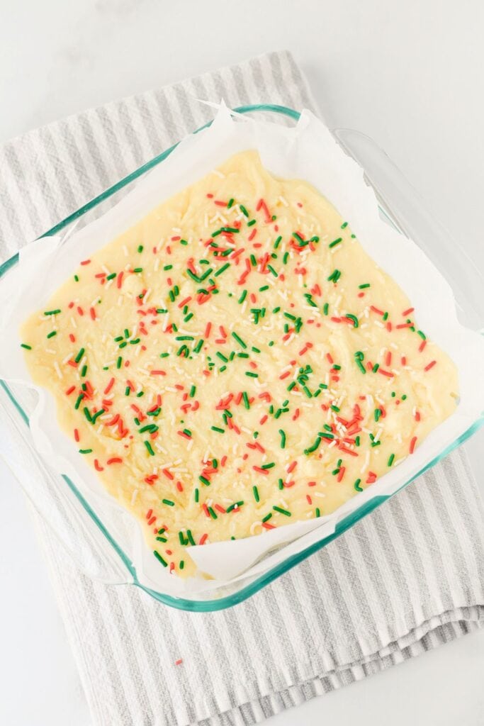 Over head view of the uncut white chocolate Christmas fudge in a glass baking dish, lined with parchment paper and topped with red, green, and white sprinkles. The fudge rests on a striped kitchen towel, ready to chill before slicing.