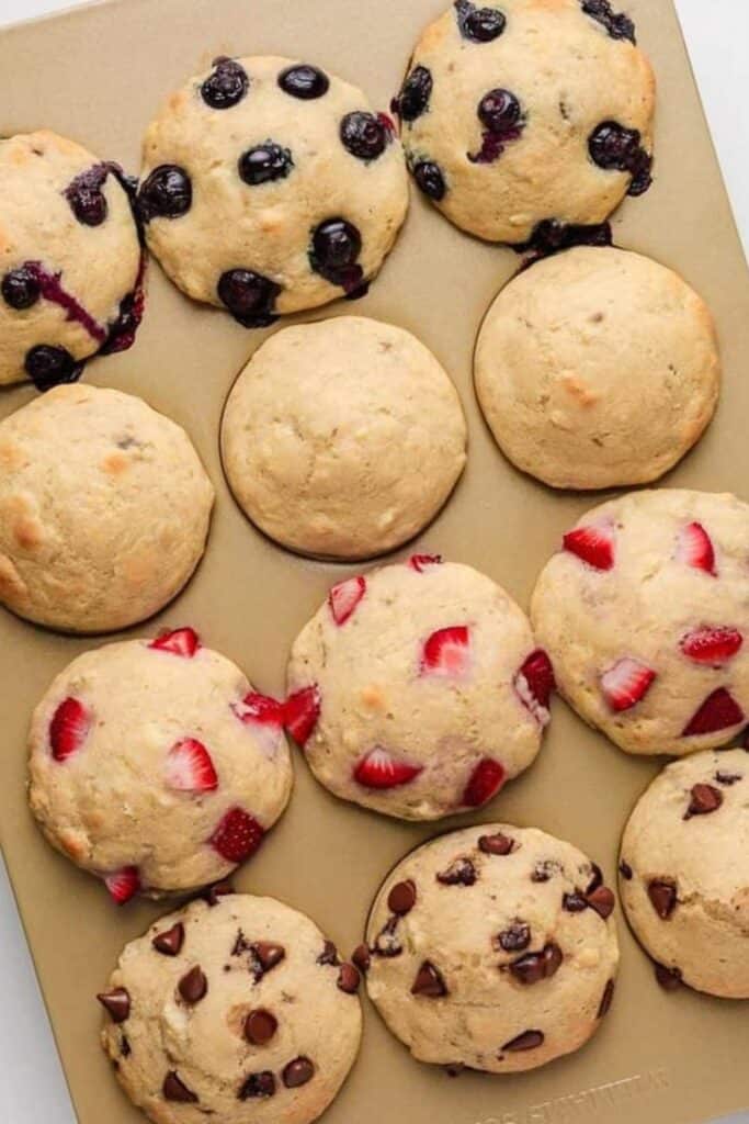 Freshly baked easy pancake muffins in a nonstick muffin tin, featuring a variety of toppings including blueberries, strawberries, chocolate chips, and plain.