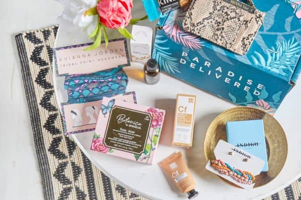 A flat lay of items from a "Paradise Delivered" travel subscription box, including skincare products, a tribal print headwrap, a faux snakeskin clutch, floral face masks, a gold bowl with colorful bracelets, and a blue envelope on a white table with a woven mat and fresh flowers. Products are arranged to showcase a relaxing, self-care travel theme.