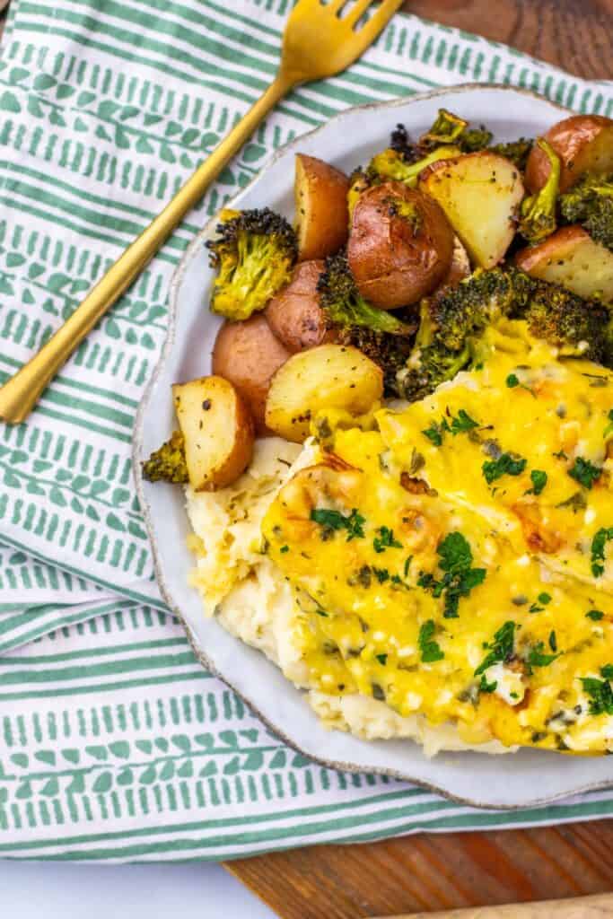 Sheet pan parmesan chicken served over creamy mashed potatoes with roasted broccoli and red potatoes on the side. The chicken is topped with melted parmesan and herbs and plated beside golden roasted vegetables for a hearty sheet pan dinner.