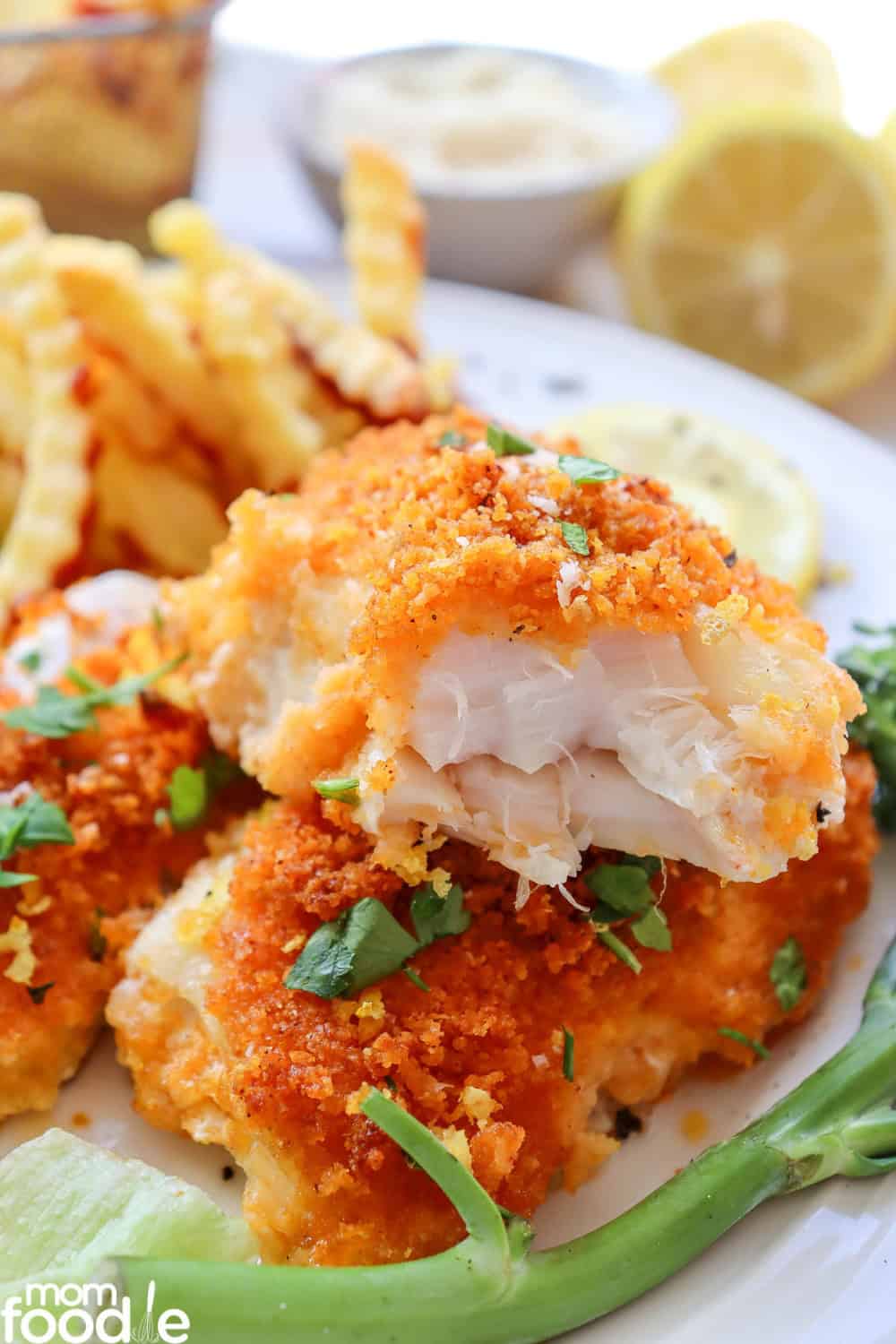 Parmesan crusted cod with a golden, crispy breadcrumb topping and flaky white fish visible in a cut piece lifted above the plate. The cod is garnished with fresh parsley and served with crinkle cut fries, lemon slices, and green vegetables for an easy seafood dinner.