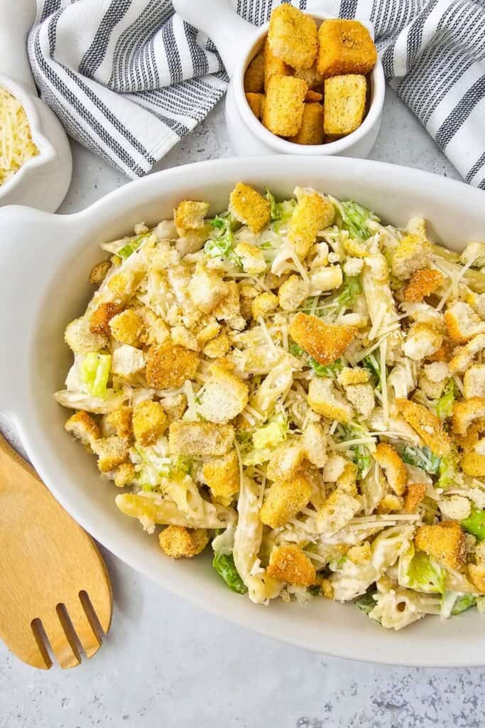 Close up of a creamy Caesar pasta salad with penne, chopped romaine, shaved Parmesan, and golden croutons in a white serving dish. A small bowl of extra croutons, shredded cheese, a wooden serving spoon, and a striped cloth surround the dish, showing an easy cold pasta salad idea ready to serve.