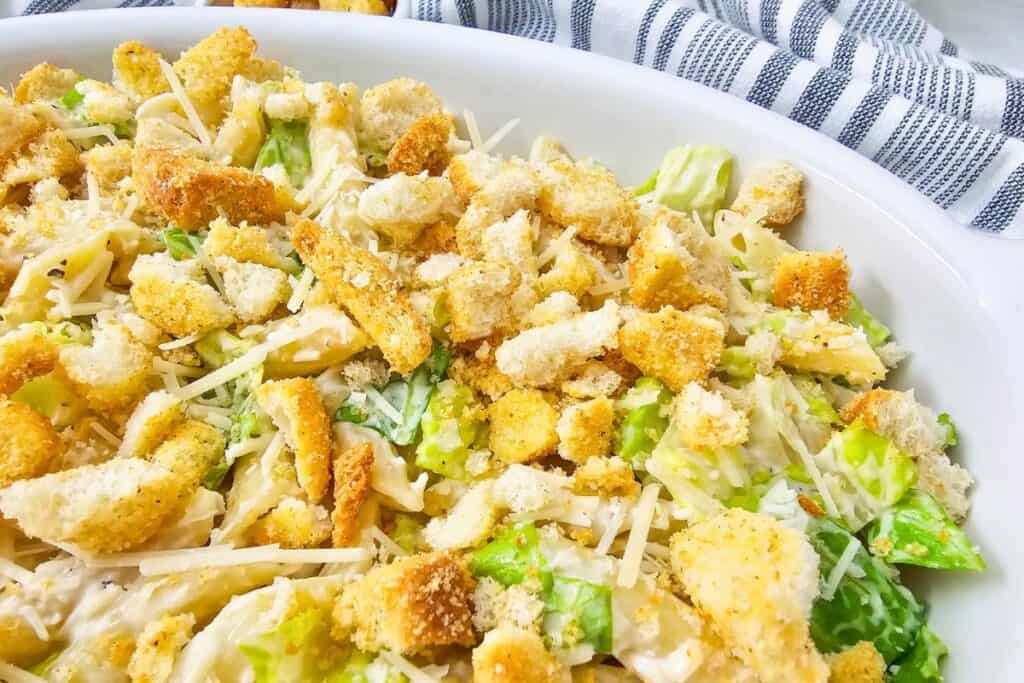 Close up of creamy Caesar pasta salad with penne, chopped romaine, shaved Parmesan, and crispy croutons in a white serving dish, showing a simple cold lunches meal prep option ready to portion and serve.
