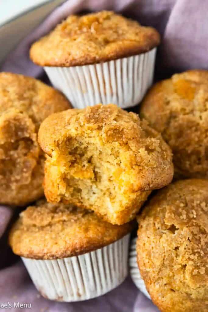 Peach muffins with golden crumbly tops and juicy peach pieces baked inside shown stacked with one broken open to reveal a soft moist interior.
