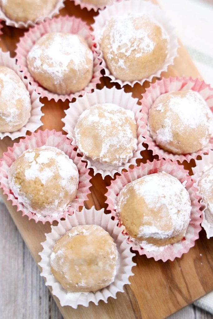 Close-up of round, powdered sugar-coated cookie balls placed in pink and white paper liners on a wooden board.