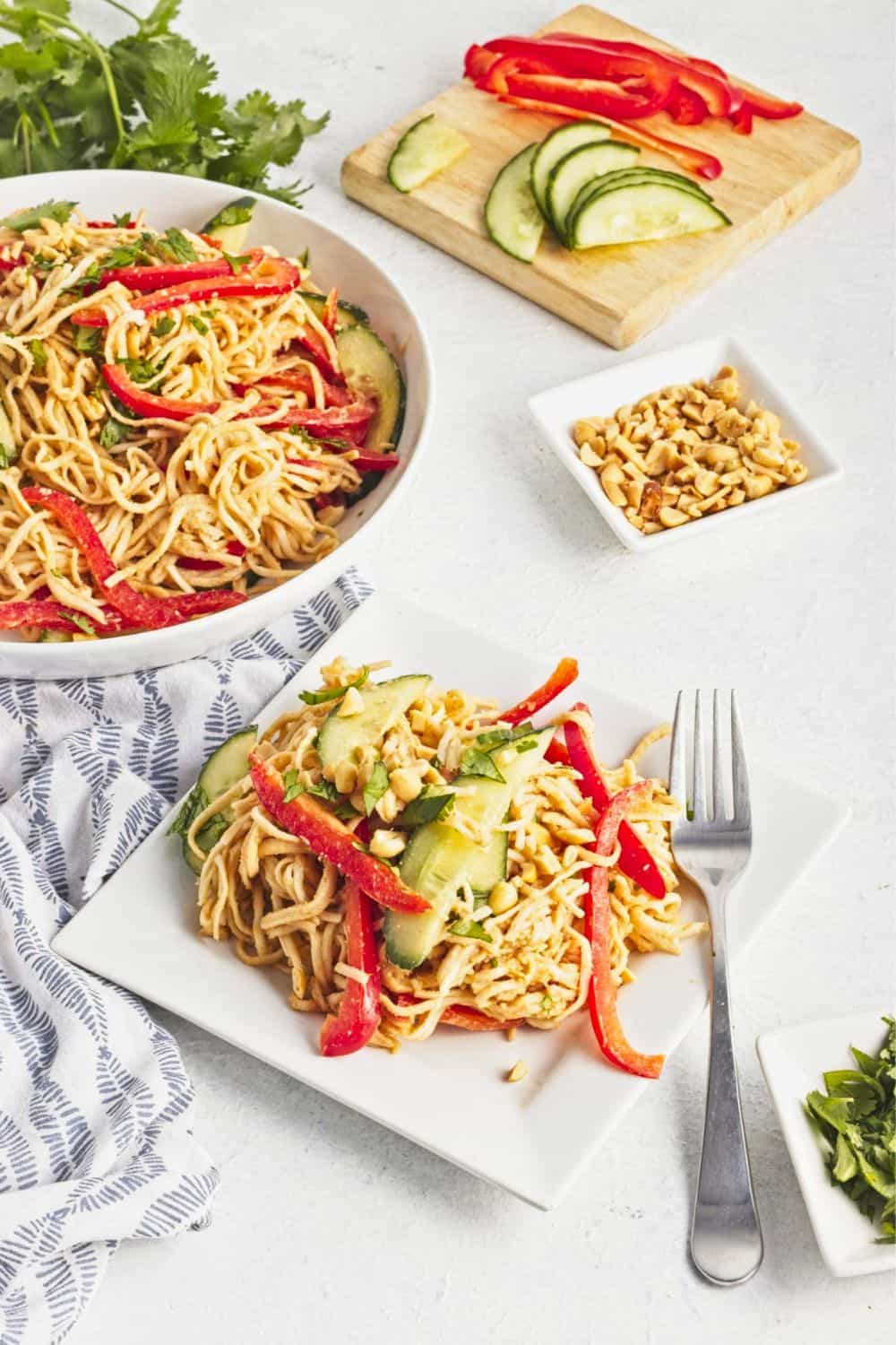 Plate of peanut butter noodles tossed with shredded chicken, sliced cucumber, red bell pepper, fresh herbs, and chopped peanuts, with a serving bowl and fresh ingredients in the background. These peanut butter noodles come together in just 20 minutes for a fresh, flavorful weeknight meal.