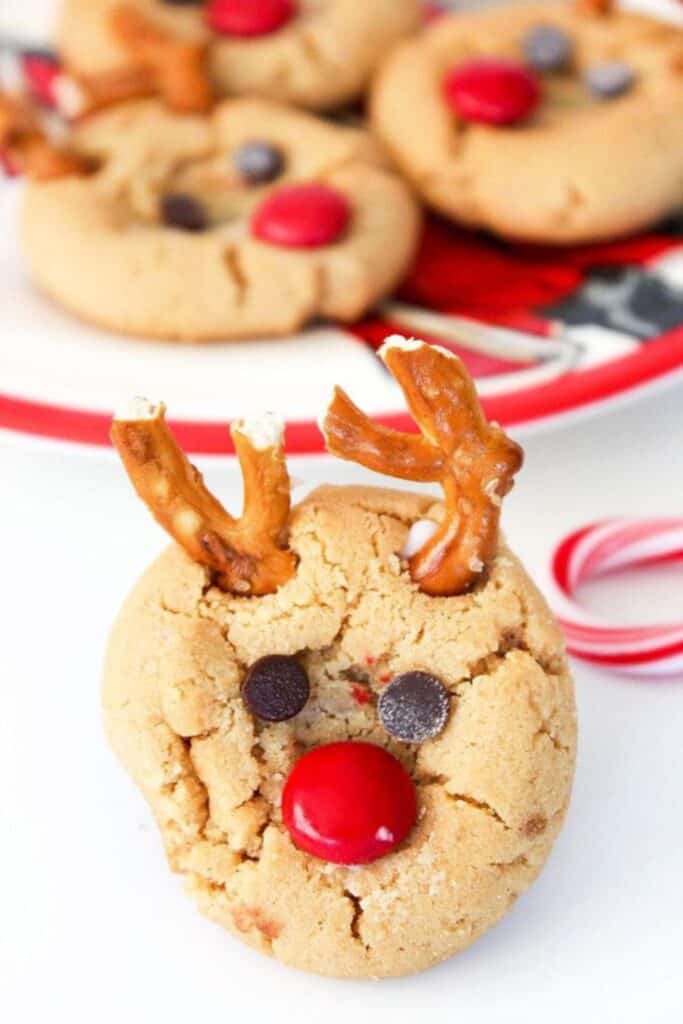 Peanut butter cookies are decorated as reindeer using chocolate chips for eyes, a red M&M for the nose, and broken pretzel twists as antlers. These peanut butter reindeer cookies are served on a holiday-themed plate with a candy cane nearby.