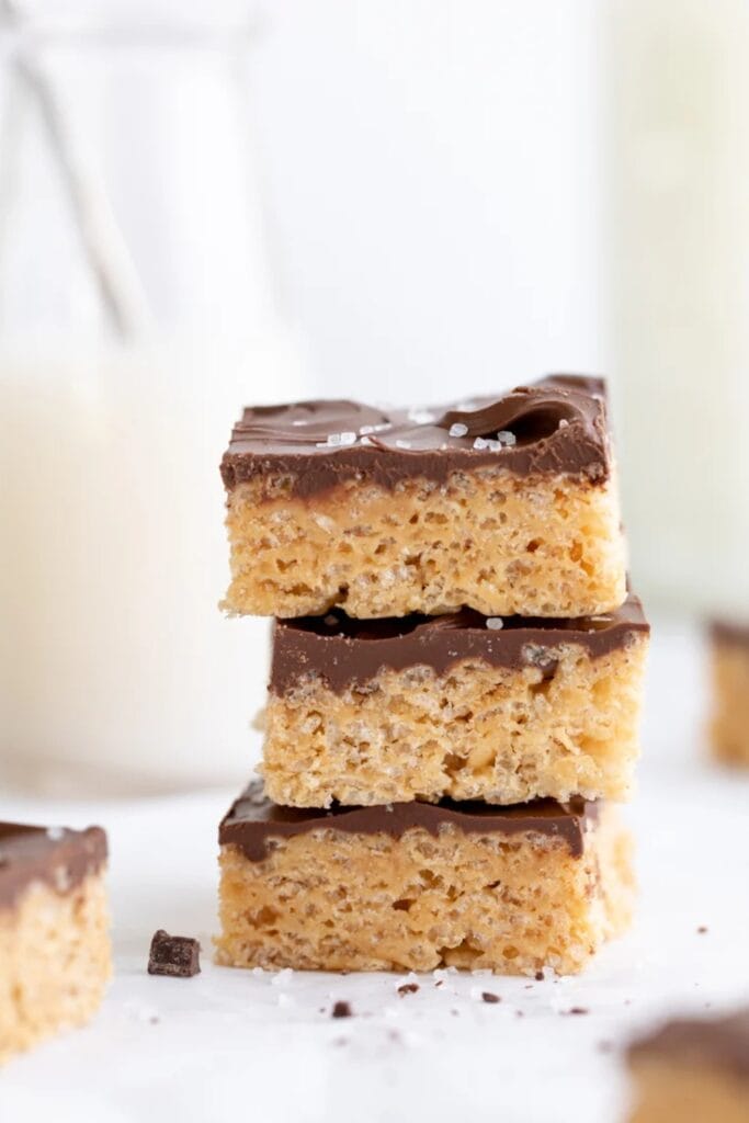 Stack of three peanut butter Rice Krispie treats topped with a smooth layer of chocolate and a sprinkle of flaky sea salt. The bars have a thick, chewy cereal base with a glossy chocolate coating on top.