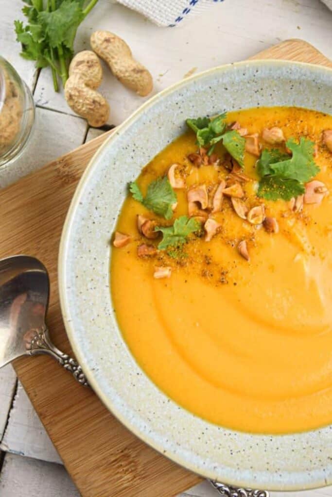 Peanut sweet potato soup is served smooth and creamy in a shallow bowl. Chopped peanuts fresh cilantro and spices top the vibrant orange soup.